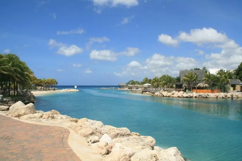 A calm, protected inlet on Curaçao: flat turquoise water between low coral-rock banks, palm trees and thatched sun shelters along the far shore