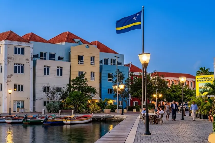 Willemstad waterfront at dusk: pale yellow and blue townhouses under red roofs, the Curaçao flag flying from a mast, lit street lamps and small wooden rowing boats moored along the quay