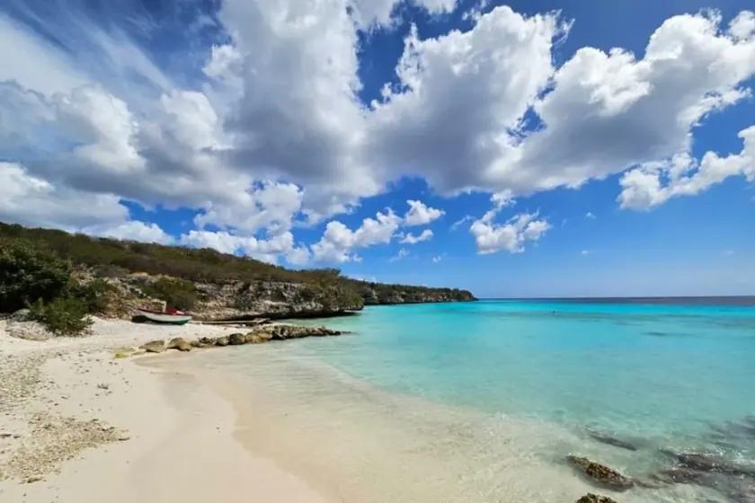 A west-coast Curaçao beach: white sand curving into shallow turquoise water, a low limestone headland covered in scrub, and tall cumulus cloud over the open sea