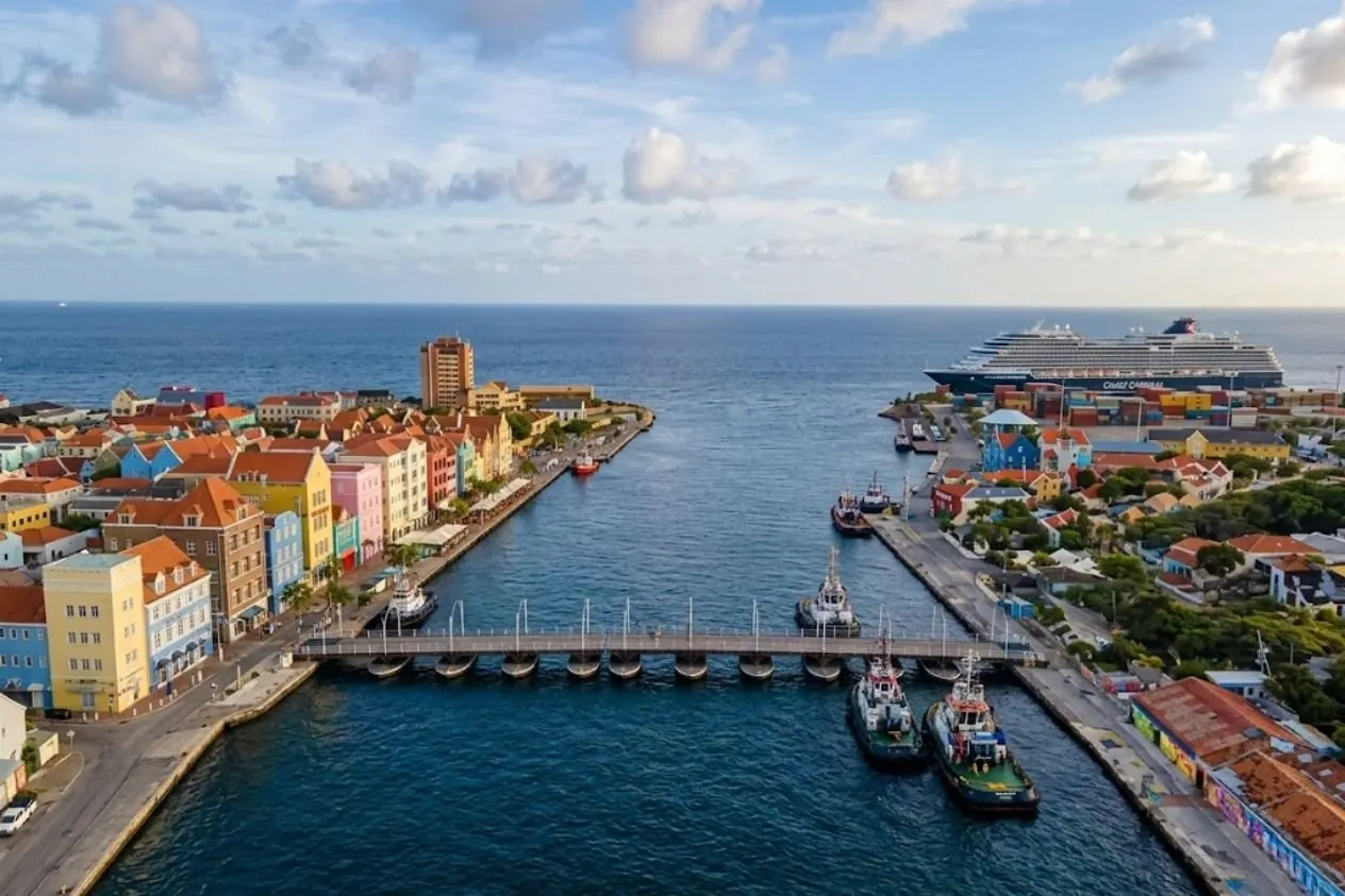 Willemstad from the air: the row of pastel merchant houses along the waterfront, the Queen Emma pontoon bridge across the harbour mouth, tugs at the quay and a cruise ship alongside the terminal